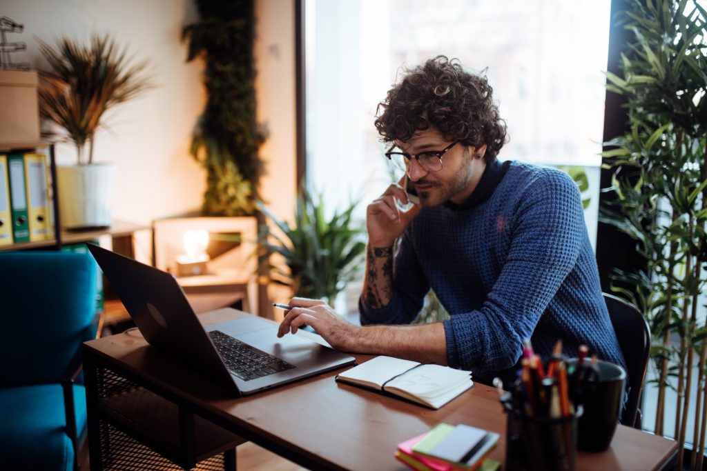 Young man talking on the phone and typing on his laptop at home